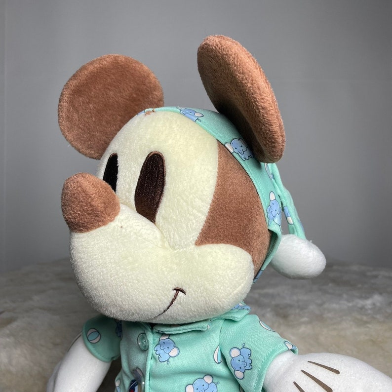 Może przedstawiać: Pluszowa zabawka Mickey Mouse w zielono-niebieskiej piżamie z wzorem słoni. Zabawka ma brązowe uszy i brązowy nos.