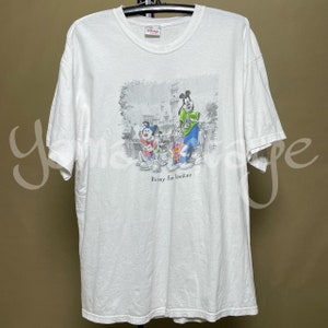 Puede incluir: Camiseta blanca con un estampado gráfico de Mickey Mouse, Goofy y Pluto frente a un castillo. El texto "Disney Fun Seeker" está impreso debajo de los personajes.