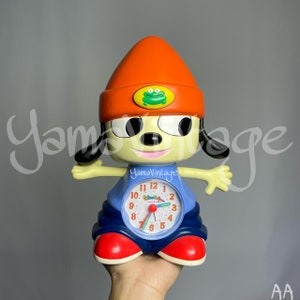 May include: A blue and orange alarm clock shaped like a cartoon dog wearing a red and orange hat. The clock face has a green frog on it and the text "Yama Vintage" is printed on the clock's body.