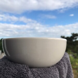 May include: A white ceramic bowl with a rounded shape, sitting on a gray towel against a blue sky background.
