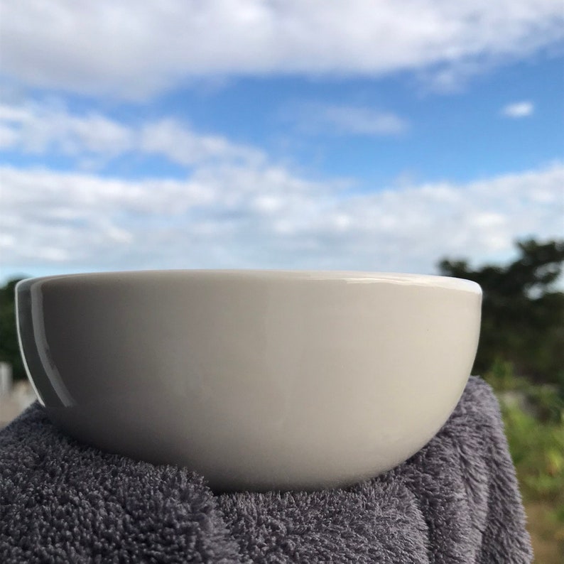 May include: A white ceramic bowl with a rounded shape, sitting on a gray towel against a blue sky background.