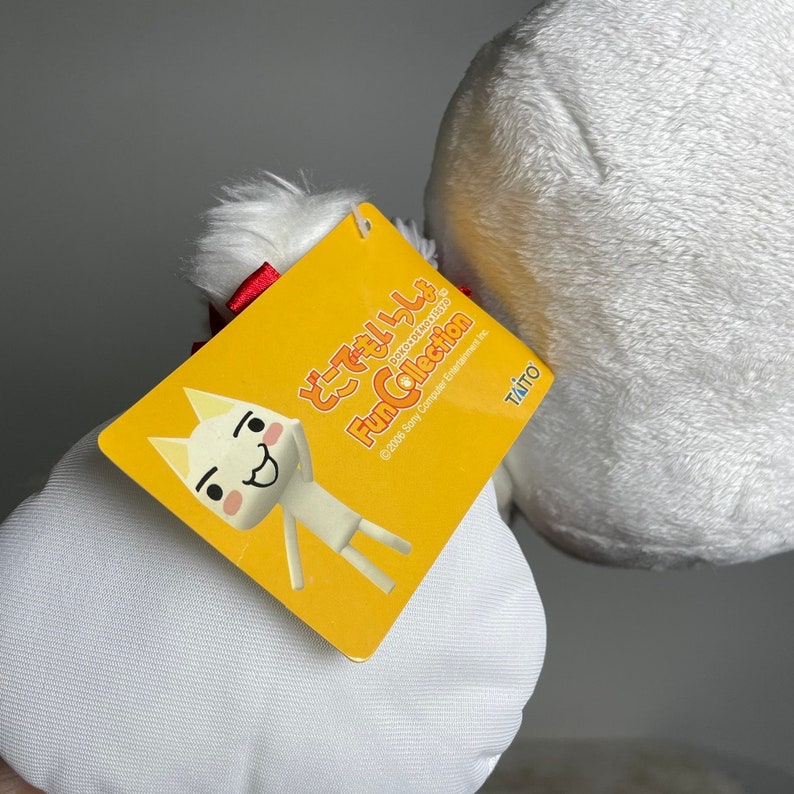 May include: A white plush toy with a red bow. The toy is labelled "Fun Collection" and features a cartoon character with a white body and a yellow face. The label also includes the text "&copy;2006 Sony Computer Entertainment Inc.  Taito"