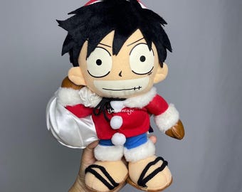 May include: Plush Luffy doll from the anime series One Piece, wearing a red jacket with white trim, blue shorts, and brown shoes. The doll has black hair, large eyes, and a stitched scar under his left eye. He is holding a white bag.
