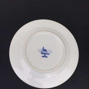 Op de afbeelding: Een wit keramiek bord met een blauw-wit logo met de tekst "PRESENTED by KONAMI" en een klein kerkje.