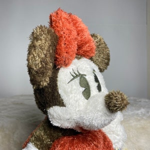 May include: A plush toy of a cartoon mouse with brown fur, a red bow, and white fur around the eyes. The mouse has embroidered eyelashes and a green eye.