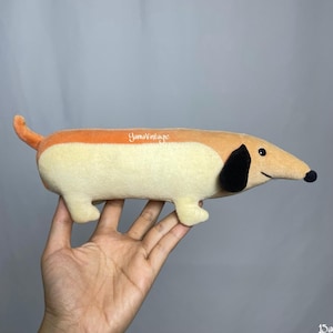 Op de afbeelding: Een pluche teckel hondenspeelgoed in tinten oranje, bruin en crème. Het speelgoed heeft een lang lichaam, korte poten en een zwart oor. De neus van de hond is zwart en hij heeft een lachende uitdrukking. Het speelgoed wordt in een hand gehouden.