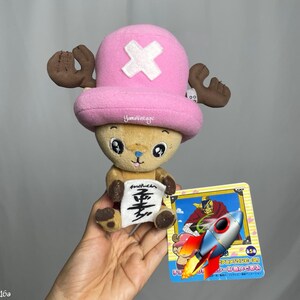 以下が含まれることがあります： アニメシリーズOne Pieceのキャラクター、チョッパーのぬいぐるみ。チョッパーは白い十字架と茶色の角が付いたピンク色の帽子をかぶっています。彼は日本語の文字が入った小さな看板を持っています。ロケットグラフィックのカードも表示されています。