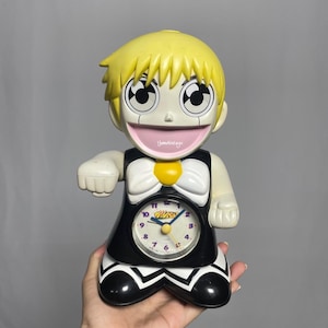 以下が含まれることがあります： 黄色い髪の漫画のキャラクターの目覚まし時計で、黒と白のドレスを着て、大きく笑っています。時計の文字盤は黒地に白の数字で、黄色と紫のデザインが施されています。