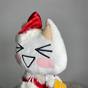 May include: White plush cat wearing a red Santa hat with a white fur trim. The cat has a smiling face with black eyes and a pink nose. The cat is wearing a red and white Santa suit.