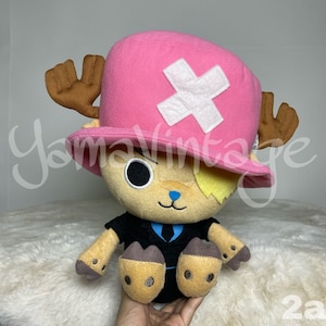 以下が含まれることがあります： アニメワンピースのトニートニー・チョッパーのぬいぐるみ。チョッパーは白い十字架のついたピンクの帽子と黒いスーツを着ています。彼は茶色の角と青い鼻をしています。