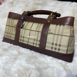 May include: A rectangular handbag with a tan plaid pattern and brown leather trim. The bag has a top handle, brass hardware, and a small tag. The bag is sitting on a white, fluffy surface. The text "YomaVintage" is visible.