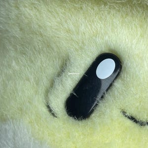 May include: Close-up of a plush toy's eye, featuring a black oval with a white circle in the center. The plush is a light yellow color.