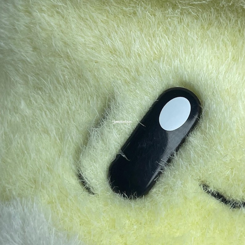 May include: Close-up of a plush toy's eye, featuring a black oval with a white circle in the center. The plush is a light yellow color.