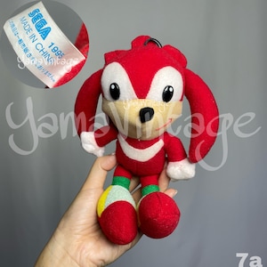 May include: A red plush toy of the character Knuckles from the Sonic the Hedgehog series. The toy has white accents, large black eyes, and a tag that reads "SEGA 1995 MADE IN CHINA". The plush is held in a hand.