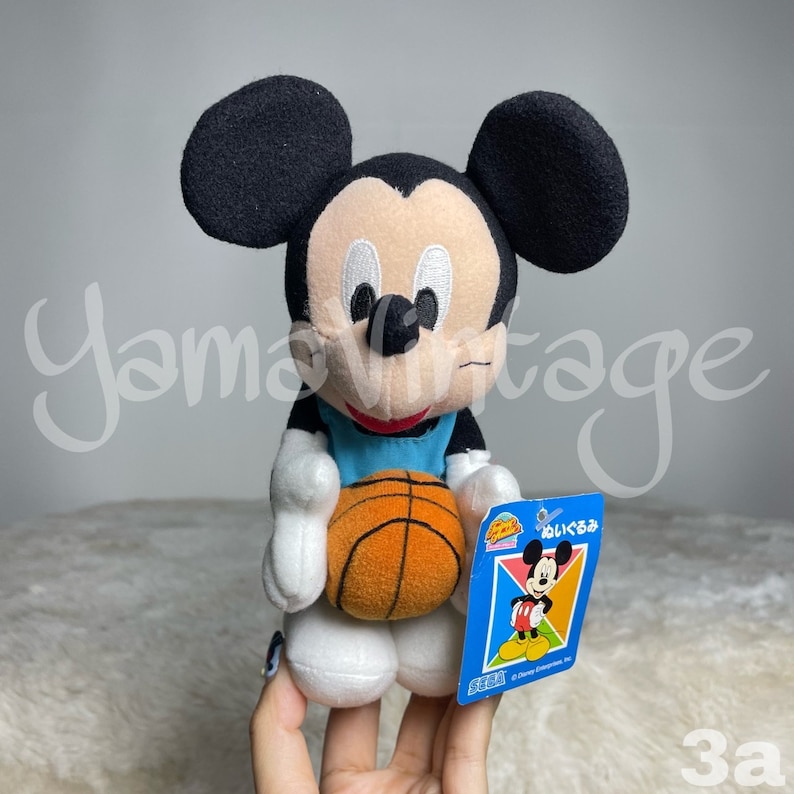 K&ouml;nnte beinhalten: Ein Pl&uuml;sch-Mickey-Mouse-Spielzeug, das ein blaues Basketballtrikot tr&auml;gt und einen orangefarbenen Basketball h&auml;lt. Das Spielzeug hat ein Etikett mit der Aufschrift "はい くま" in Japanisch.
