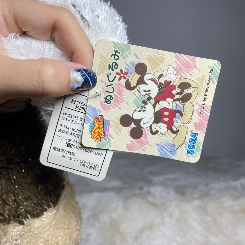 May include: A plush toy of Mickey Mouse and Minnie Mouse hugging. The toy is brown and white with black eyes and red cheeks. The tag attached to the toy reads "Fantasy Amase" and "ぬいぐるみ" in Japanese. The tag also includes contact information for Sega and Disney Enterprises, Inc.