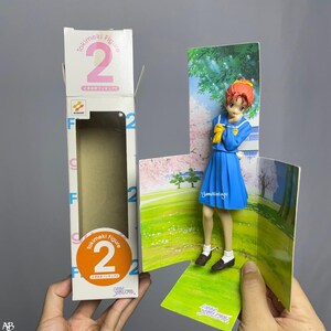 Peut inclure: Une figurine d'anime bleue et blanche représentant une fille aux cheveux roux portant un uniforme scolaire. La figurine est posée sur un présentoir en carton avec un fond rose et vert. La figurine tient un foulard jaune et porte un uniforme scolaire bleu et blanc. La figurine fait partie d'une série de figurines Tokimeki.