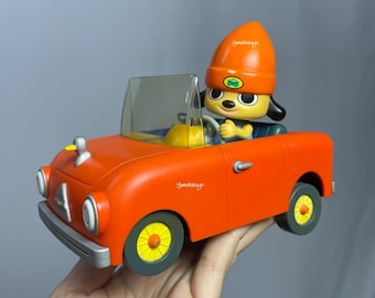May include: A red toy car with a yellow steering wheel and a yellow and green dog character wearing a red and orange hat driving the car.