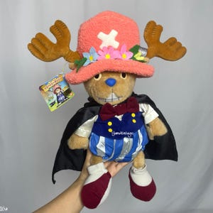 以下が含まれることがあります： アニメ「ワンピース」のキャラクターのぬいぐるみ。茶色のクマのぬいぐるみで、白い十字架が付いたピンク色の帽子、茶色の角、青と白の衣装を着ています。黒いマントとタグが付いています。