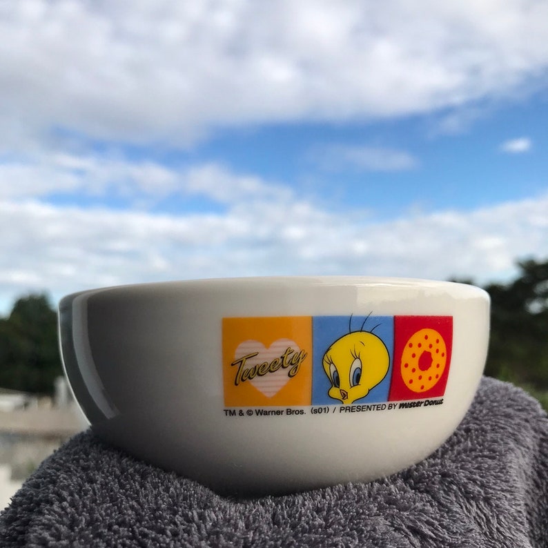 May include: White ceramic bowl with a Tweety Bird design. The bowl features a yellow heart with the word "Tweety" in pink lettering, a blue square with Tweety Bird, and a red square with a donut. The text "TM & &copy; Warner Bros. (s01) / PRESENTED BY MASTER DOLLS" is printed on the bottom of the bowl.