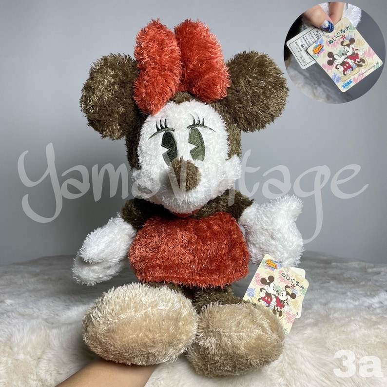 May include: A plush brown and white Minnie Mouse toy wearing a red dress and a red bow. The toy has a brown nose and large black eyes. The toy is holding a tag that says "Minnie Mouse" in Japanese.