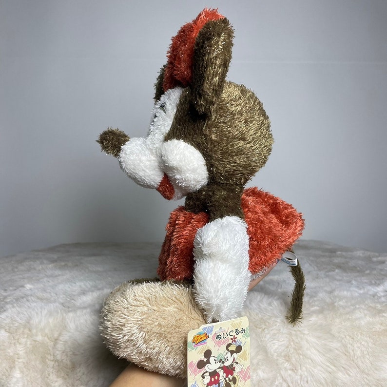 May include: A brown and white plush toy mouse wearing a red and white dress. The mouse is sitting with its tail curled behind it. The toy has a tag that says "あいうえお" in Japanese.