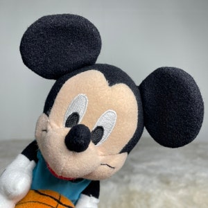 K&ouml;nnte beinhalten: Ein Pl&uuml;sch-Mickey-Mouse-Spielzeug, das ein blaues Trikot tr&auml;gt und einen orangefarbenen Basketball h&auml;lt. Das Spielzeug hat schwarze Ohren, eine schwarze Nase und gestickte Augen.