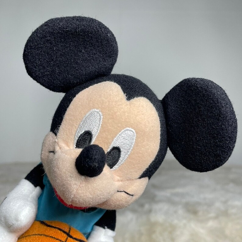 K&ouml;nnte beinhalten: Ein Pl&uuml;sch-Mickey-Mouse-Spielzeug, das ein blaues Trikot tr&auml;gt und einen orangefarbenen Basketball h&auml;lt. Das Spielzeug hat schwarze Ohren, eine schwarze Nase und gestickte Augen.