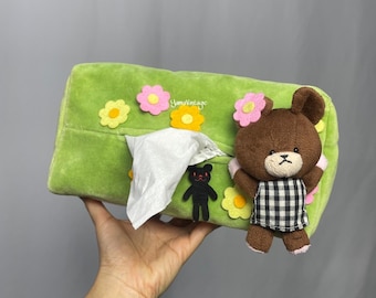 May include: Green plush tissue box cover with embroidered flowers and a brown bear wearing a checked dress. The bear is holding a black bear with red eyes.