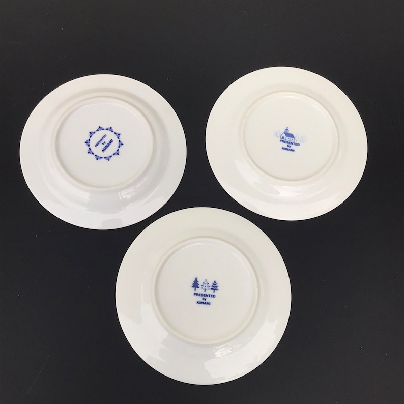 Op de afbeelding: Drie witte keramische borden met blauwe en witte ontwerpen. Elk bord heeft een ander ontwerp, waaronder een kerk, een stervorm en een groep bomen. De borden zijn allemaal gemarkeerd met "Presented by Holland".