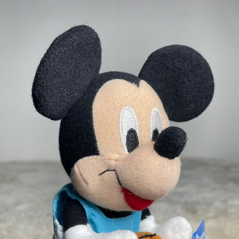 K&ouml;nnte beinhalten: Ein Pl&uuml;sch-Mickey-Mouse-Spielzeug, das ein blaues Basketballtrikot tr&auml;gt. Er hat ein breites L&auml;cheln und h&auml;lt einen Basketball.