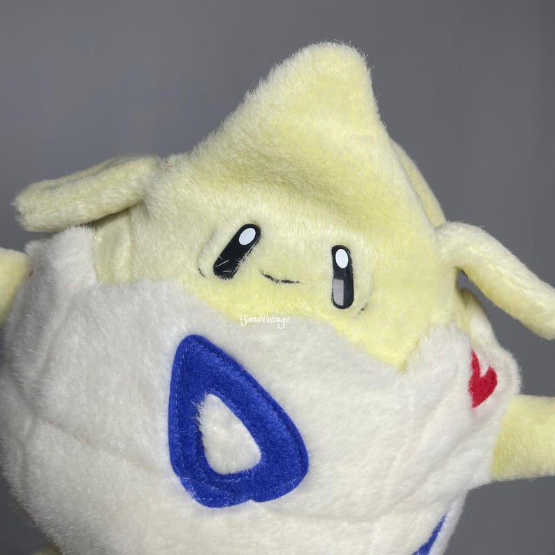 May include: A plush toy of a white and yellow Pokemon character with blue and red accents. The character has a large, round head with a pointed top and a small, round body. The character has large, black eyes and a small, pink mouth.