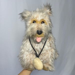 Può includere: Un cane di peluche con occhi marroni, una lingua rosa che sporge e un cordoncino nero con un'etichetta in pelle marrone intorno al collo. Il cane è marrone chiaro e bianco con una texture di pelliccia irsuta.