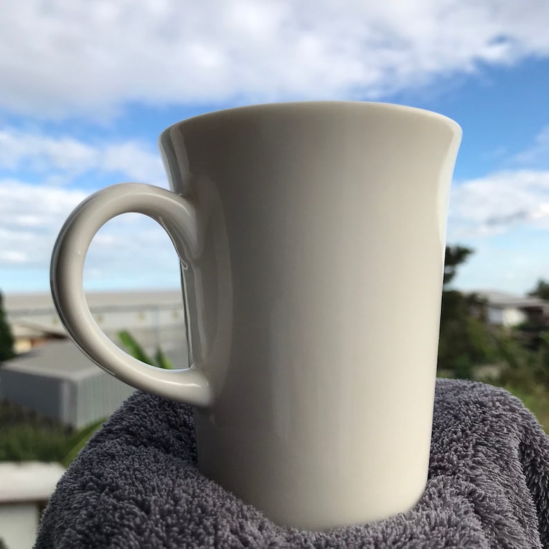 May include: A white ceramic mug with a handle, sitting on a gray towel. The mug is empty and the background is a blue sky with white clouds.