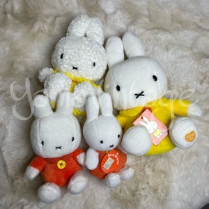 Könnte beinhalten: Vier Plüsch-Hasen aus weißem Plüsch mit unterschiedlichen Kleidungsfarben. Ein Hase trägt ein gelbes Hemd, ein anderer ein orangefarbenes Hemd und zwei tragen gelbe Hemden mit dem Text "Dick Bruna" darauf. Die Hasen-Plüschtiere sitzen auf einer weißen, flauschigen Oberfläche.