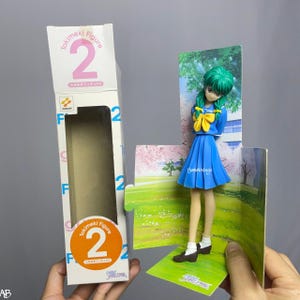 Peut inclure: Figurine d'anime Tokimeki Figure 2 dans une boîte. La figurine représente une fille aux cheveux verts, portant un uniforme scolaire bleu et des chaussures marron. La boîte est blanche avec le numéro 2 et les mots "Tokimeki Figure".