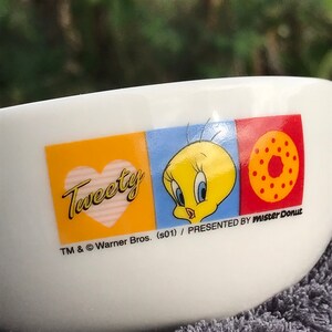 May include: White ceramic bowl with a Tweety Bird design. The bowl has a yellow, blue, and red background with a heart, Tweety Bird, and a donut. The text "Tweety" is in a yellow heart with pink stripes. The text "TM & &copy; Warner Bros. (s01) / PRESENTED BY Mister Donut" is at the bottom of the bowl.