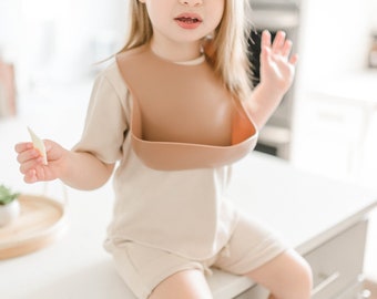 Puede incluir: Un niño pequeño con una camisa beige de manga corta y un babero de silicona marrón se sienta en una encimera blanca. El niño sostiene un pequeño trozo de comida en su mano.