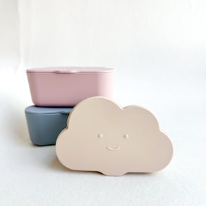 May include: Three stackable containers in pink, gray, and beige. The beige container is shaped like a cloud with a smiling face.