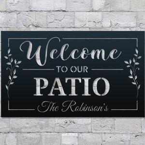 以下が含まれることがあります： 黒い金属製の看板に白い文字で「Welcome to our Patio The Robinson's」と書かれています。看板は、両側に葉のついたつるが装飾的に描かれた枠で囲まれています。