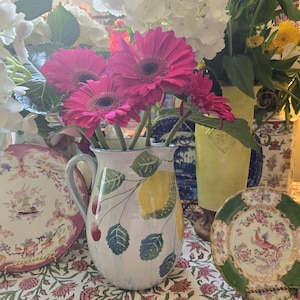 May include: A white ceramic pitcher with a yellow lemon and green leaves design. The pitcher is filled with pink gerbera daisies and white hydrangeas.
