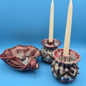 Peut inclure: Un ensemble de trois bougeoirs en céramique en forme de coquillage. Les bougeoirs sont décorés d'un glaçage rose, blanc et violet et ont des accents dorés. L'ensemble comprend deux bougeoirs et un petit plat.