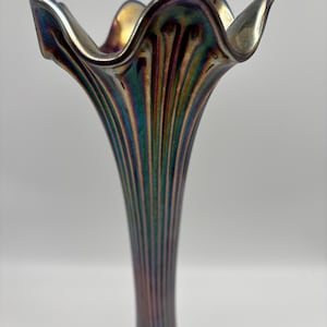 May include: An iridescent art glass vase with a flared, ruffled rim. The vase features vertical ribbing and a color-shifting finish, displaying hues of gold, green, and purple. The vase has a slender, elongated shape.