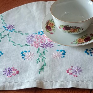 May include: White embroidered tea cozy with a floral design in shades of pink, blue, and green. A white teacup and saucer with floral designs are on top of the cozy.
