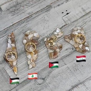 May include: Four phone ring holders shaped like the countries of Israel, Lebanon, Jordan, and Iraq. Each holder is made of a clear resin with embedded seashells and a gold-colored ring. The holders are displayed on a woodgrain surface with flags of the countries below them.