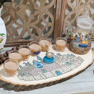 May include: A wooden serving tray with a painted cityscape design featuring a mosque and buildings. Six clear glass cups filled with a light brown liquid are arranged around the tray. A glass teapot with a painted cityscape design is on the tray.