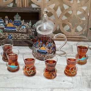 May include: A set of six clear glass teacups with intricate hand-painted designs, including a mosque, a prayer rug, and a rosary. The teacups are filled with a light brown liquid. A matching glass teapot with a similar design is also included.