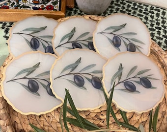 May include: Set of six white coasters with a gold rim and a printed design of olive branches and olives. The coasters are arranged in a circle on a woven basket.