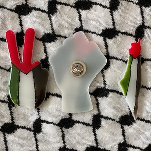May include: Three Palestinian flag pins, each with a different design: a peace sign, a fist, and an outline of Palestine. The pins are made of resin and have a silver pin back.