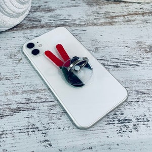 May include: A white smartphone with a black and red peace sign phone ring holder attached to the back. The ring holder has a silver metal ring.